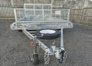 Utility trailer with a metal mesh cargo area and a spare tire mounted on the hitch, parked on gravel with a wooden fence in the background.