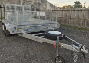 Aluminum utility trailer with a mesh cargo cage on a gravel lot, spare tire mounted on the hitch.