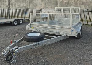 Metal trailer with mesh cage and spare tire mounted on the hitch, parked on gravel with a larger trailer in background.
