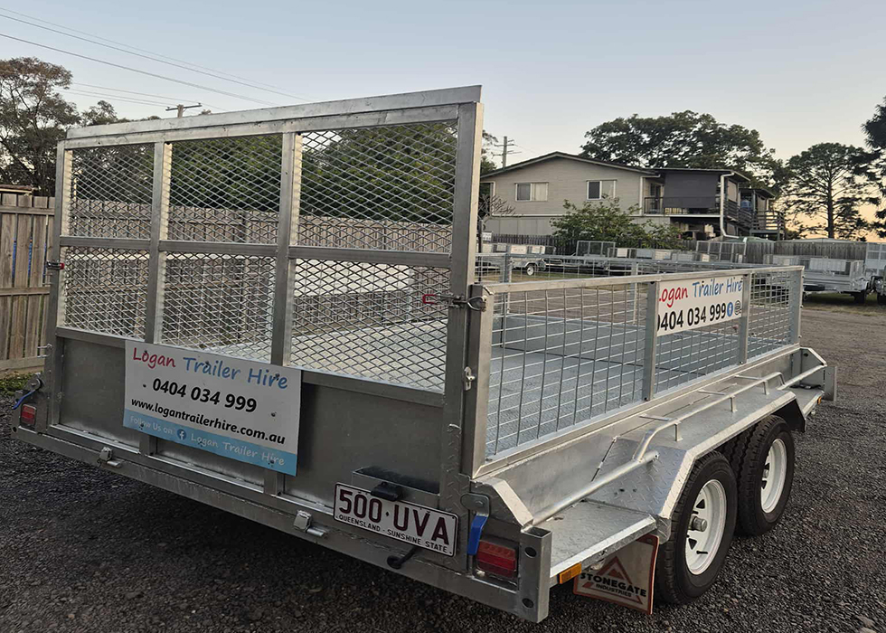 Dual-axle flatbed trailer with metal mesh sides and gate, advertised for hire by Logan Trailer Hire in a gravel lot at dusk.