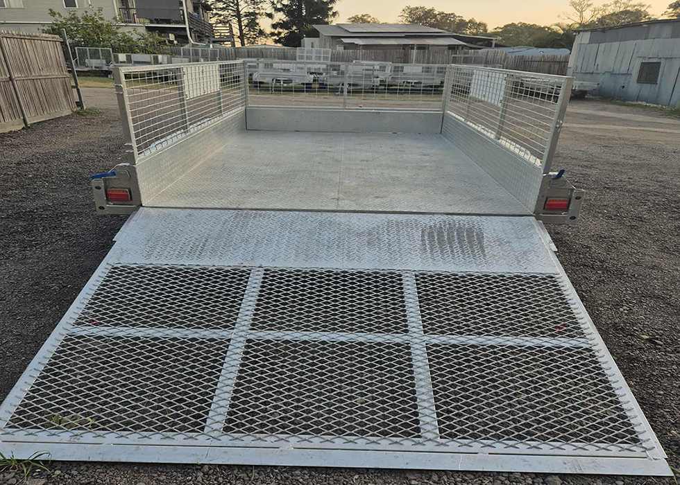 Aluminum utility trailer with mesh sides and a lowered rear ramp, parked on a gravel lot in a yard.