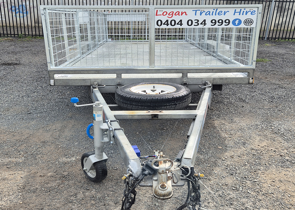 Metal utility trailer on gravel with mesh sides and a spare wheel mounted on the frame; sign reads 'Logan Trailer Hire' with phone number on the back.