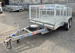 Galvanized dual-axle trailer with a mesh cargo cage and spare tire, parked on a gravel lot with a Logan Trailer Hire sign nearby