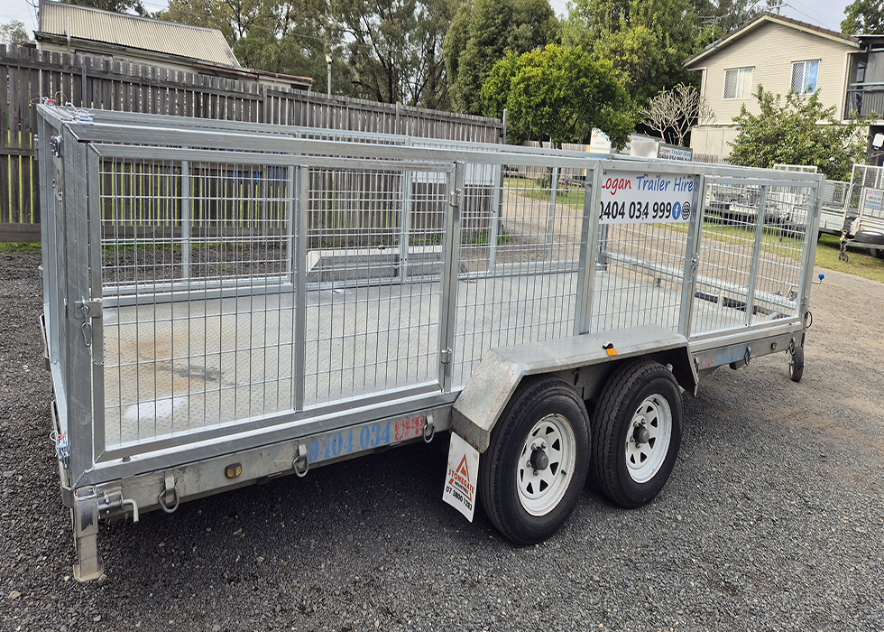 Silver metal utility trailer with mesh sides and a double axle, parked on a gravel lot; has a sign for Logan Trailer Hire on the side.