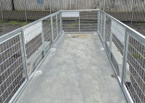 Metal grated walkway with wire-mesh railings and a gate at the far end, outdoors beside a tall fence